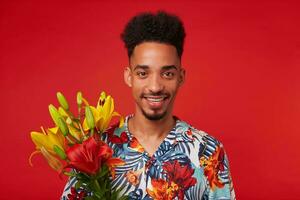 cerca arriba alegre joven africano americano chico, usa en hawaiano camisa, mira a el cámara con contento expresión, sostiene amarillo y rojo flores, soportes terminado rojo antecedentes. foto