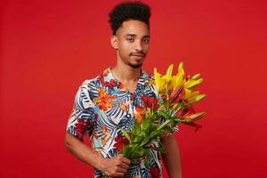 Portrait of cheerful young African American man, wears in Hawaiian shirt, looks at the camera and smile, holds yellow and red flowers, stands over red background. photo