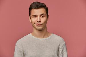 Portrait of handsome young man with raised eyebrow in disapproval, wears in blank t-shirt, looks at the camera with a grin and doubts, stands over pink background. photo