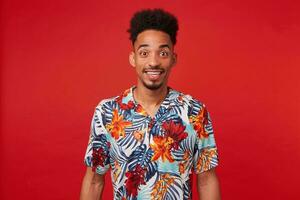 retrato de contento joven africano americano chico, usa en hawaiano camisa, mira a el cámara con alegre expresión, soportes terminado rojo antecedentes y en general sonrisas foto
