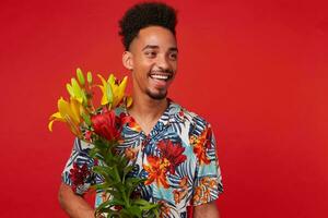 Portrait of laughing young African American guy, wears in Hawaiian shirt, looks at the camera with happy expression, holds yellow and red flowers, stands over red background. photo