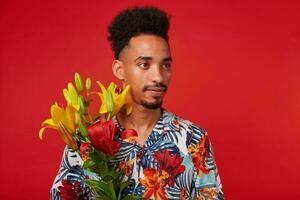 Calm young African American man, wears in Hawaiian shirt, looks away, holds yellow and red flowers, stands over red background. photo