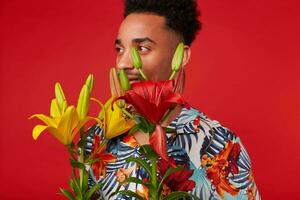 Close up of young African American guy, wears in Hawaiian shirt, looks away, stands over red background, yellow and red flowers covered his face. photo