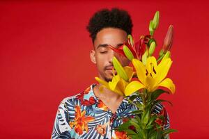 Portrait of young calming African American man, wears in Hawaiian shirt, holds yellow and red flowers bouquet and covered face, stands over red background with closed eyes. photo