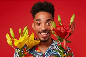 Close up cheerful young African American guy, wears in Hawaiian shirt, looks at the camera with happy expression, stands over red background, yellow flowers covered face. photo