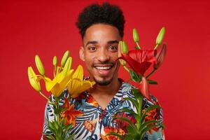 Close up cheerful young African American guy, wears in Hawaiian shirt, looks at the camera with happy expression, holds yellow and red flowers, stands over red background. photo