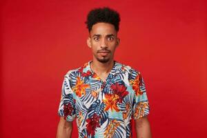 Young wondered African American man, wears in Hawaiian shirt, looks at the camera with surprised expression and wide opened eyes, stands over red background. photo