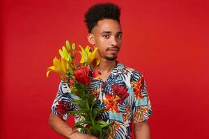 retrato de calma joven africano americano chico, usa en hawaiano camisa, mira a el cámara , sostiene amarillo y rojo flores, soportes terminado rojo antecedentes. foto