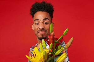 Close up young dark skinned man, wears in Hawaiian shirt, looks at yellow and red flowers with disgust, he doesn't like the smell, stands over red background. photo