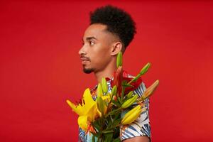 Portrait of young calm African American man, wears in Hawaiian shirt, holds yellow and red flowers bouquet, stands over red background. photo