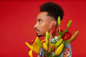 Close up of young calm African American man in Hawaiian shirt, holds yellow and red flowers bouquet, stands over red background. photo