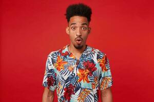 Young surprised African American man, wears in Hawaiian shirt, looks at the camera with amazed expression and wide opened eyes, stands over red background. photo