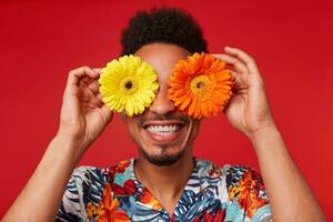 cerca arriba sonriente joven africano americano chico, usa en hawaiano camisa, mira a el cámara canal flores con contento expresión, sostiene amarillo y rojo flores, soportes terminado rojo antecedentes. foto