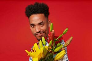 Close up young cool dark skinned man, wears in Hawaiian shirt, looks at the camera with funny expression, holds yellow and red flowers, stands over red background. photo