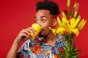 cerca arriba de joven sorprendido africano americano hombre en hawaiano camisa, mira lejos y Bebiendo agua desde un amarillo vaso, sostiene amarillo y rojo flores ramo, soportes terminado rojo antecedentes. foto