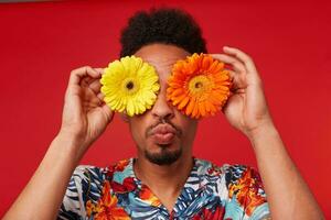 Close up positive young African American guy, wears in Hawaiian shirt, looks at the camera trough flowers with happy expression, send kiss at the camera, stands over red background. photo