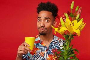 Close up of young confused African American man in Hawaiian shirt, looks at the camera and holds a yellow glass, holds yellow and red flowers bouquet, stands over red background. photo