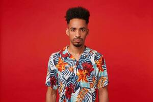 Portrait of frowning young African American guy, wears in Hawaiian shirt, looks at the camera with unhappy expression, stands over red background. photo
