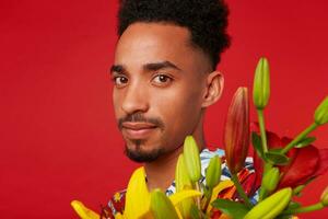 Close up of young calm dark skinned man, wears in Hawaiian shirt, holds yellow and red flowers bouquet, stands over red background and looks at the camera. photo