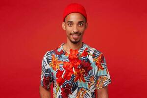 retrato de joven sonriente oscuro desollado chico, usa en hawaiano camisa y rojo sombrero, mira a el cámara, con un rojo flor, soportes terminado rojo antecedentes. foto