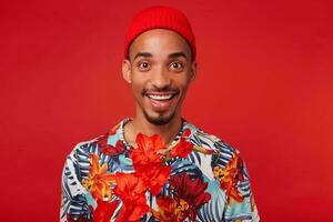 Portrait of young happy dark skinned guy, wears in Hawaiian shirt and red hat, looks at the camera with happy expression, stands over red background. photo