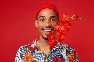 foto de joven atractivo contento oscuro desollado hombre, usa en hawaiano camisa y rojo sombrero, mira a el cámara con contento expresión, soportes terminado rojo antecedentes.