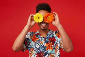 Portrait of cheerful young African American guy, wears in Hawaiian shirt, looks at the camera trough flowers with happy expression, stands over red background. photo