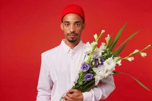 Portrait of young calm African American man, wears in white shirt and red hat, looks at the camera and holds bouquet , stands over red background. photo
