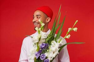 foto de joven positivo oscuro desollado hombre, usa en blanco camisa y rojo sombrero, mira lejos y sonriente, sostiene ramo de flores , soportes terminado rojo antecedentes.