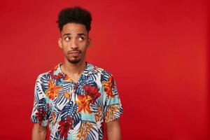 Portrait of thinking young African American guy, wears in Hawaiian shirt, looking right at copyspace, standing over red background. photo