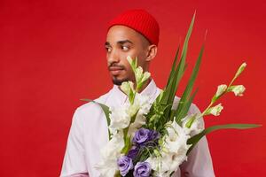 Photo of young calm dark skinned man, wears in white shirt and red hat, looks away and holds bouquet , stands over red background.