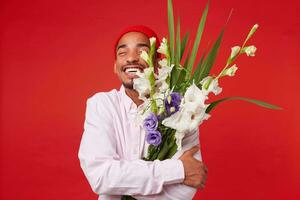 Portrait of young happy dark skinned man, wears in white shirt and red hat, with closed eyes hugs bouquet , stands over red background and smiling. photo