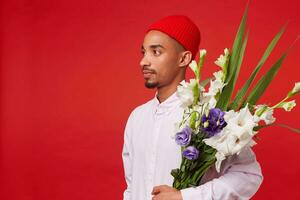 Young calm African American guy in white shirt and red hat, looks away and holds bouquet , stands over red background. photo