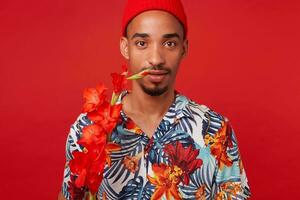 Portrait of young dark skinned guy, wears in Hawaiian shirt and red hat, looks at the camera with calm expression, with a red flower, stands over red background. photo