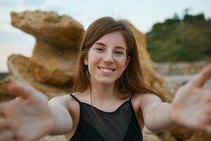 Close up of young cheerful ginger cute lady with freckles on the beach, broadly smiling and pulling hands to the camera, looks positive and happy. photo