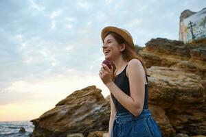 Young laughing ginger freckles lady on the beach, wears hat, eating a peach, broadly smiles and enjoy the morning on the seaside, looks positive and happy. photo