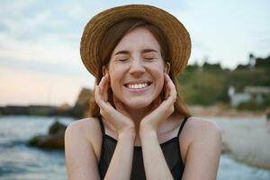 Close up of young ginger cute freckles woman walks on the beach, wears hat, touches cheeks, broadly smiles with closed eyes, looks positive and happy. photo