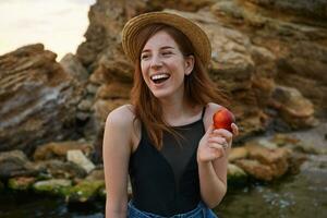 Portrait of young ginger nice freckles lady on the beach, wears hat, eating a peach, broadly smiles, looks positive and happy, laughing and looks away. photo