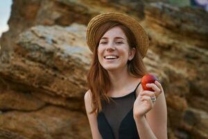 Portrait of young ginger nice freckles lady on the beach, wears hat, eating a peach, broadly smiles and looks at the camera, looks positive and happy. photo