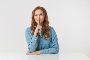 Photo of young mysterious cute blonde woman wears in denim shirts, sitting at the white table, smiling and shows silence gesture, please keep calm. Stands over white background.