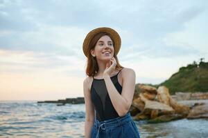 Photo of young ginger cute lady with freckles walks on the beach, wears hat, dreamily looks away and touches chees, looks positive and happy.