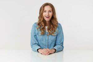 Portrait of young happy amazed cute curly blonde lady in denim shirts sitting at the white table with wide opened mouth, isolated over white background. photo