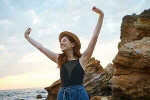 joven linda jengibre niña con pecas usa en en sombrero, en general sonrisas, mira lejos y disfrutar el Mañana en el playa ingenio elevado manos, mira alegre y contento. foto