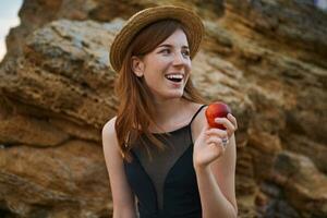 Portrait of young ginger cute freckles woman on the beach, wears hat, eating a peach, broadly smiles and looks away, looks positive and happy. photo