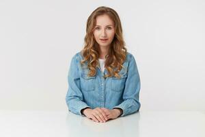 Portrait of young positive cute curly blonde lady in denim shirts sitting at the white table and smile, isolated over white background. photo