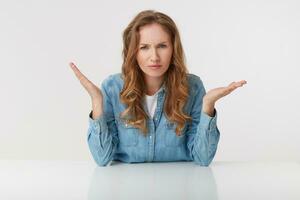 Photo of young outraged blonde woman wears in denim shirts, sitting at the white table and spreads his arms to the side, frowning and looks discontented, isolated over white background.