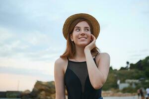 Portrait of young ginger cute freckles woman walks on the beach, wears hat, dreamily looks away and touches cheek, looks positive and happy. photo