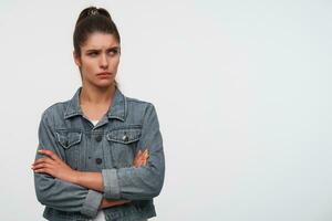 Portrait of young frown brunette lady wears in white t-shirt and denim jackets, looks away with disgusted expression, stands over white background with crossed arms. photo