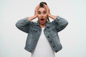 Young shocked brunette lady wears in white t-shirt and denim jackets, looks at the camera with wide open mouth and eyes, surprised expression, keep palms on the head, stands over white background. photo