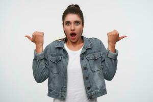 Young wondered brunette lady wears in white t-shirt and denim jackets, looks at the camera with shocked expression, points fingers to the different side, stands over white background. photo